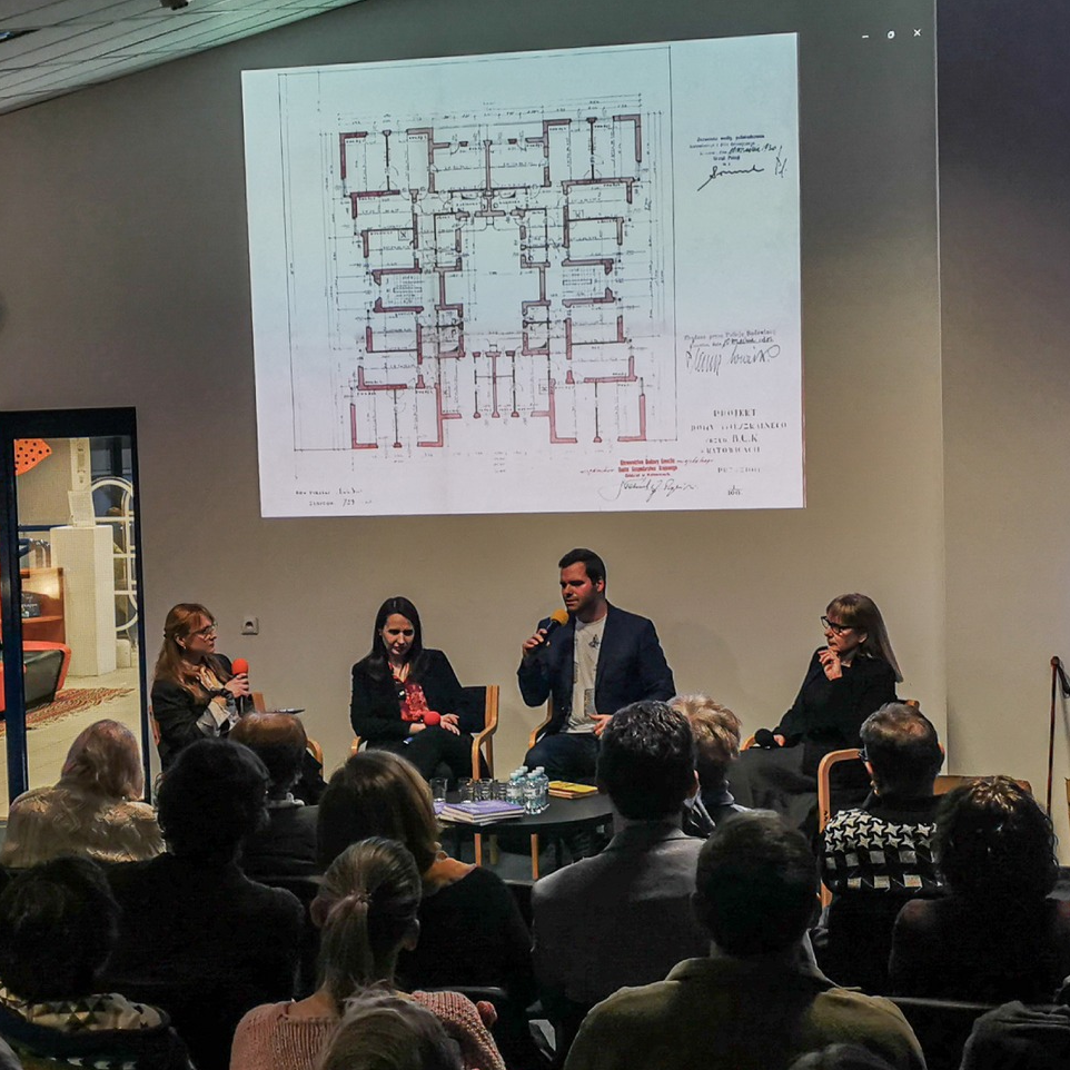 Widok z tyłu sali na prelegentów, w tle rzut budynku z książki