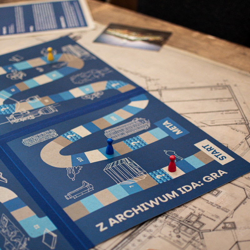 Zdjęcie przedstawia grę planszową Z ARCHIWUM IDA:GRA w tle są projekty i pocztówki, na planszy są pionki